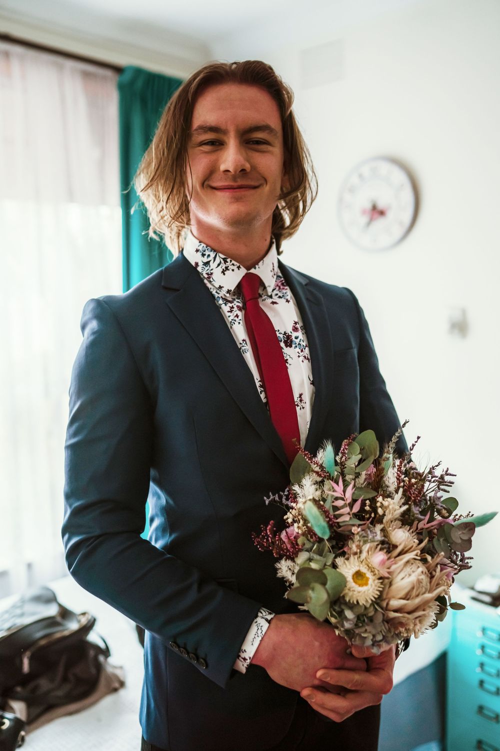 A person in a dark suit and red tie holds a rustic wedding bouquet with gerbera daisies and mixed flowers indoors.