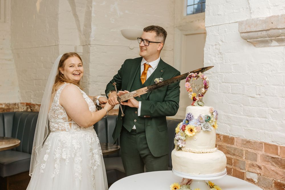 A couple cuts into their elegant three-tiered white wedding cake adorned with colorful floral decorations.