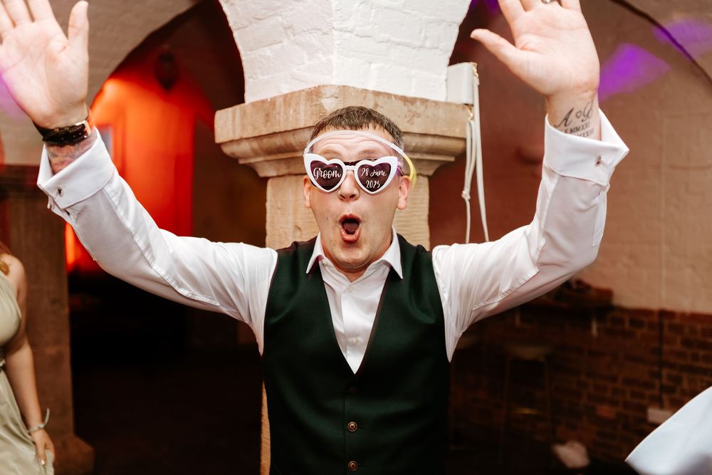 Person in sunglasses and green vest raises arms excitedly with an animated expression at a party.