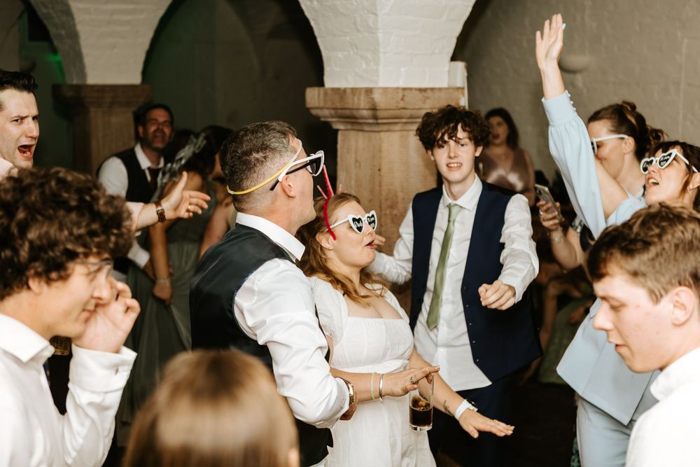 People dancing energetically in low lighting at a wedding reception in a dimly lit stone-walled venue.