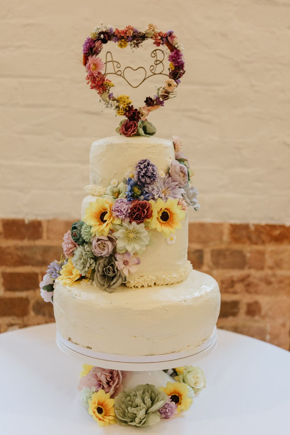 Three-tier white wedding cake decorated with colorful fresh flowers and a floral heart-shaped topper on display against brick wall.