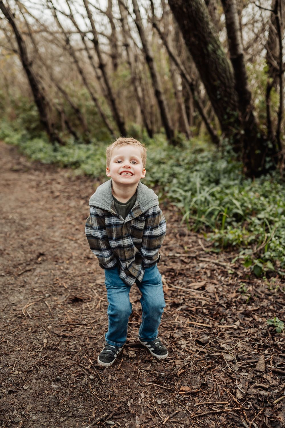 A child wearing a plaid shirt and jeans crouches in a wooded area with fallen leaves on the ground.