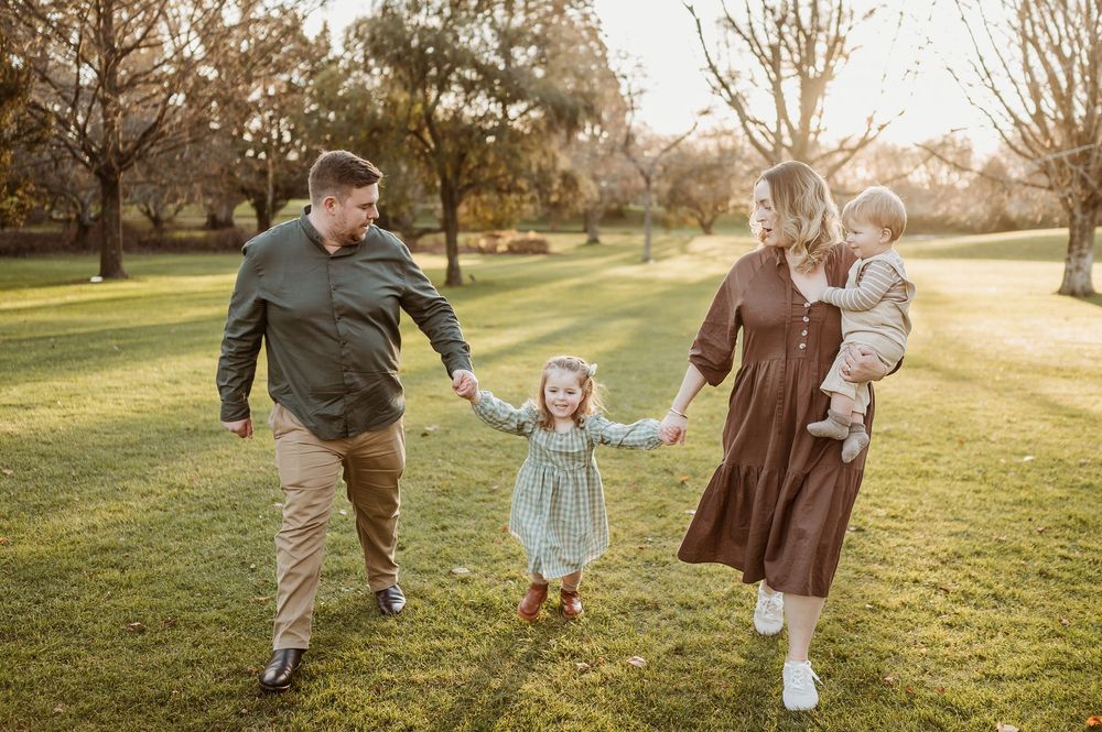 A family playfully swings a small child while walking in a sunlit park during golden hour sunset.