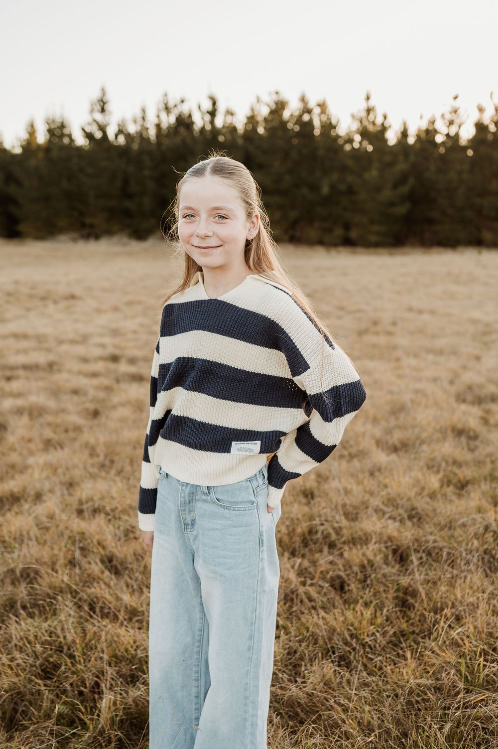 Person in light and navy striped top and light denim jeans stands in a golden field at sunset.
