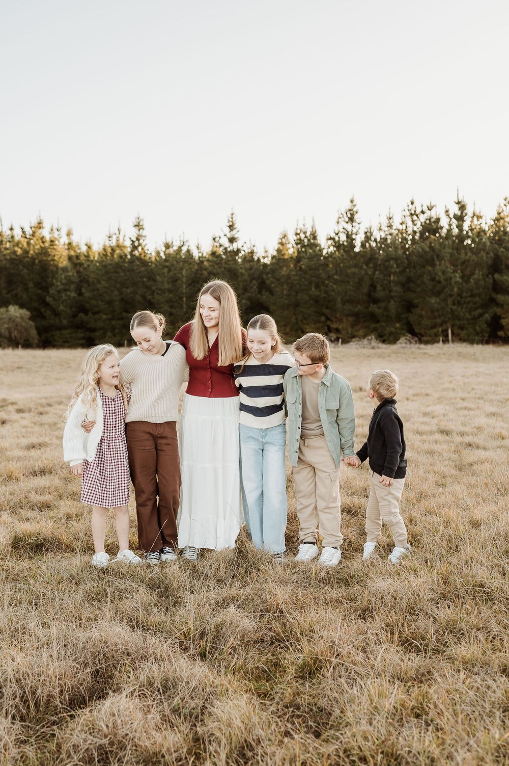 Family members stand in a golden field at sunset wearing coordinated neutral-toned outfits while holding hands and facing away.