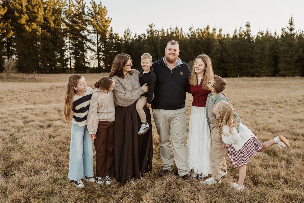 Group stands together in golden sunlit field with evergreen forest backdrop during sunset session.