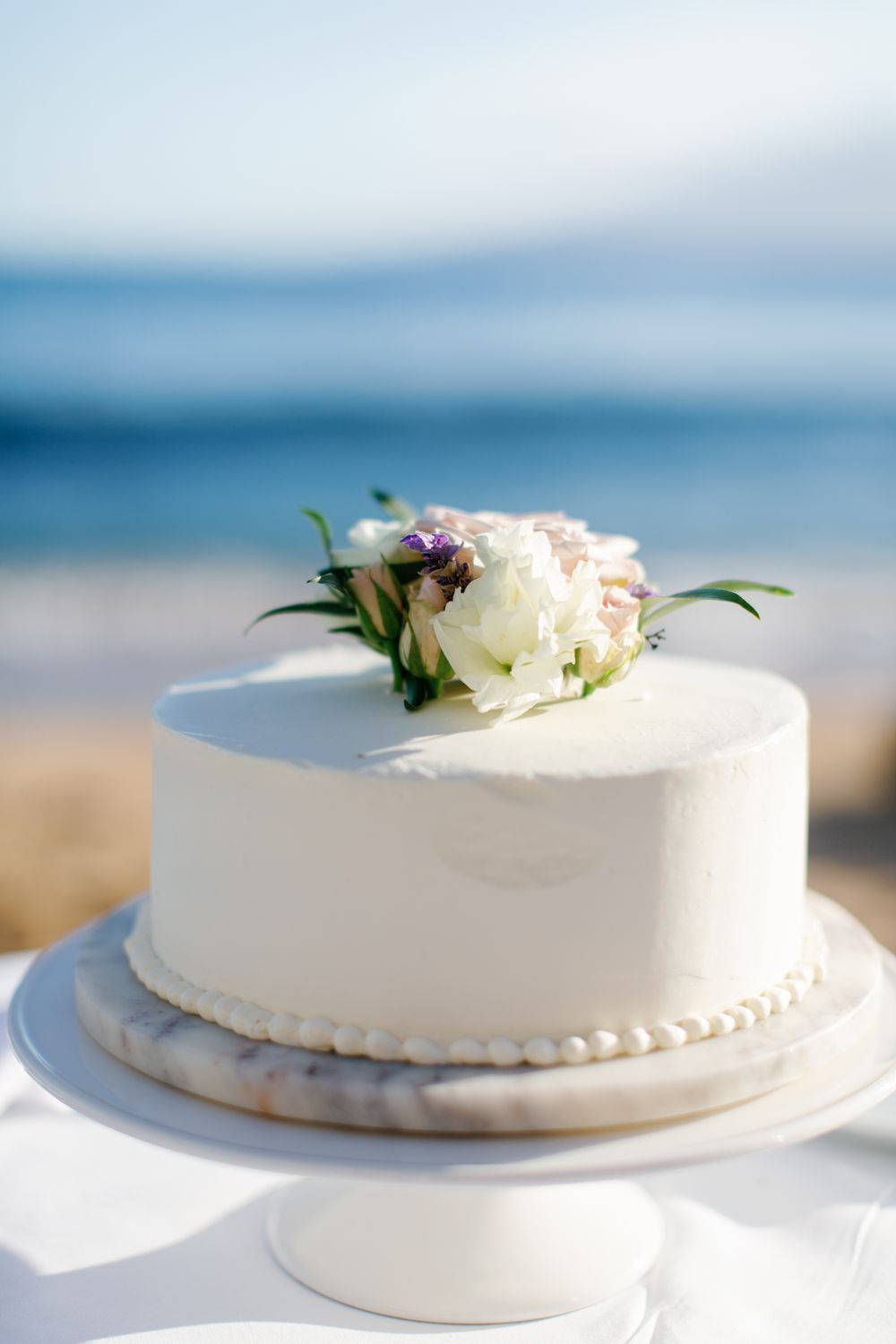 A simple white wedding cake topped with fresh white and purple flowers sits on a cake stand against a blurry beach background.