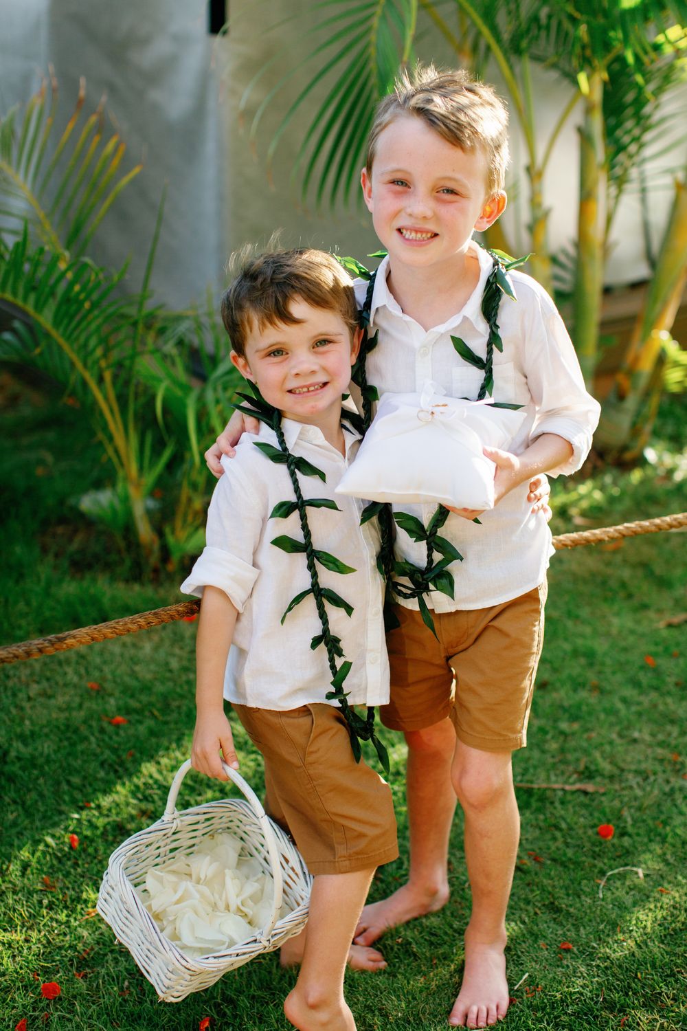 Two ring bearers in white shirts and brown shorts carry decorative baskets on a grassy lawn.