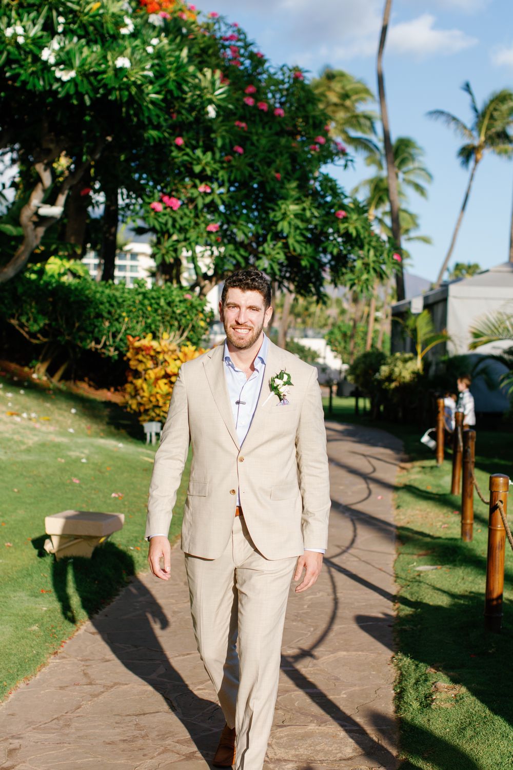 Someone walks down a tropical garden path wearing a tan linen suit with palm trees and colorful flowers in the background.