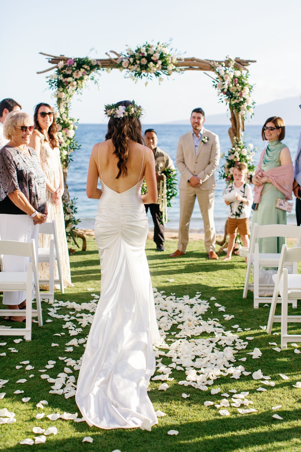 Bride in white dress walks down a beachside aisle lined with rose petals and white chairs during outdoor wedding ceremony.