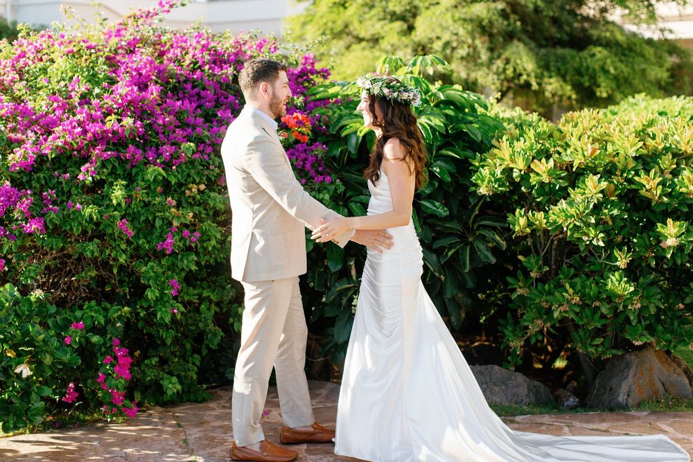 A bride and groom hold hands in front of vibrant pink bougainvillea flowers and lush greenery in a garden setting.