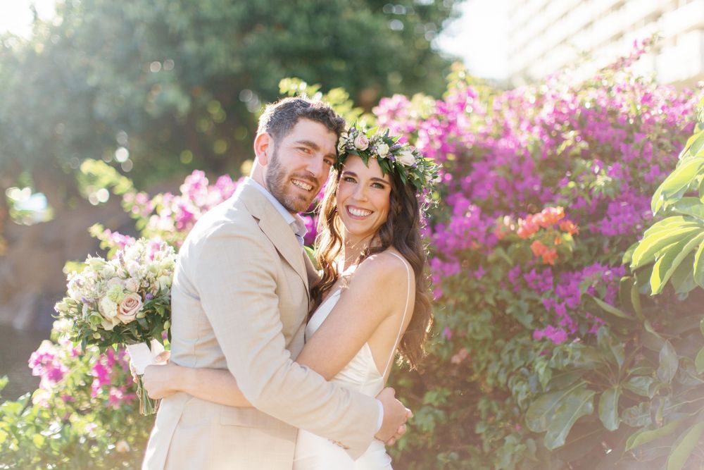 A joyful couple in wedding attire embrace outdoors with pink bougainvillea flowers and sunlight in the background.