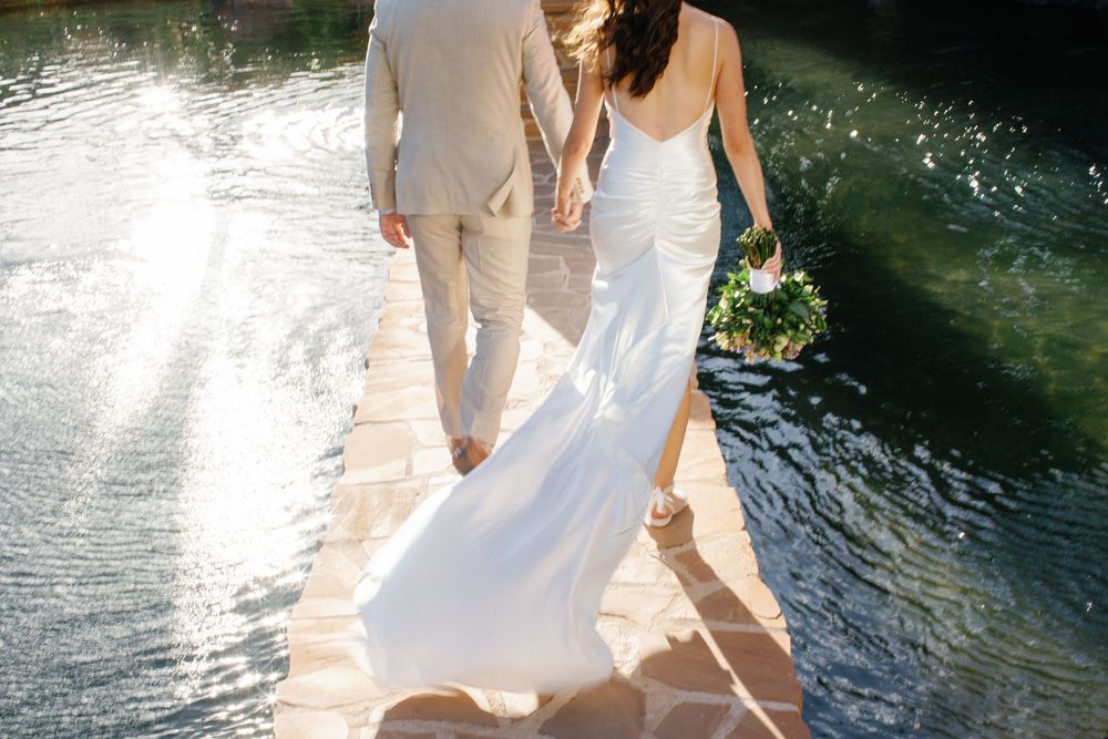 Woman in a flowing white sundress sitting by serene water's edge at sunset with rippling reflections.