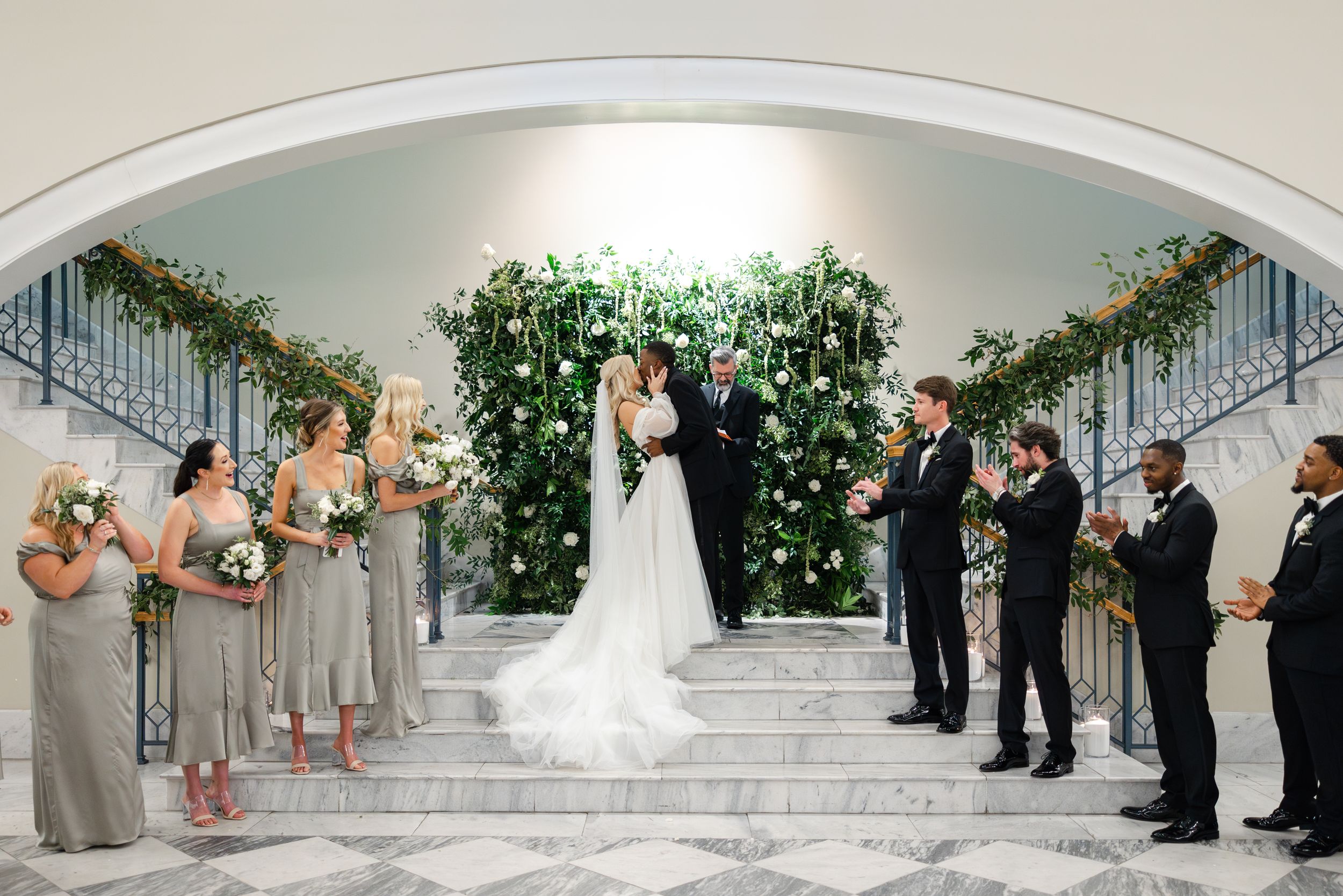 A wedding party stands symmetrically on white marble stairs with bridesmaids in gray and groomsmen in black suits.