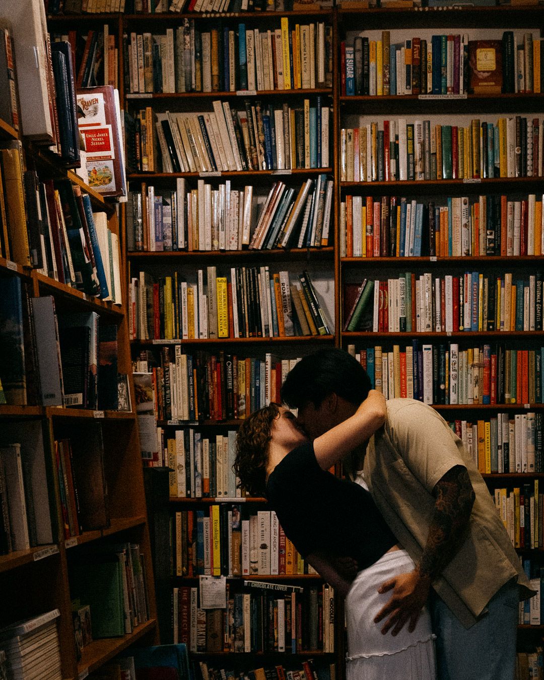 A couple kissing at the end of an aisle in a bookstore.