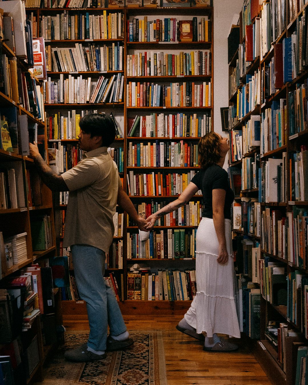 A couple browse through tall wooden bookshelves while holding hands.
