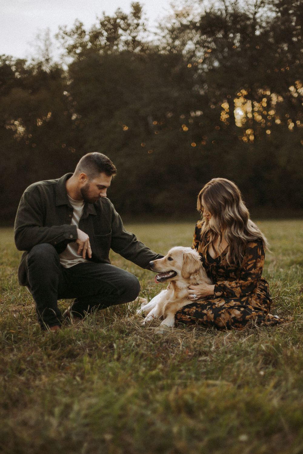 A couple sits in a golden field at sunset with their friendly golden retriever enjoying a tender moment outdoors.