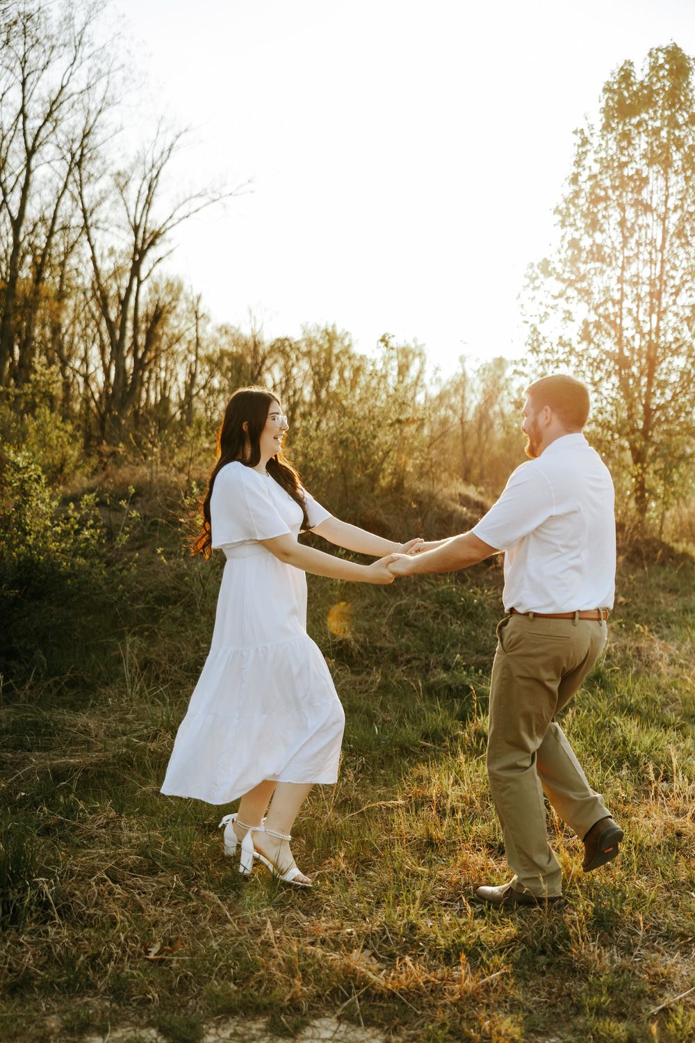 A couple dances together in a sun-drenched field during golden hour while wearing coordinating white outfits.