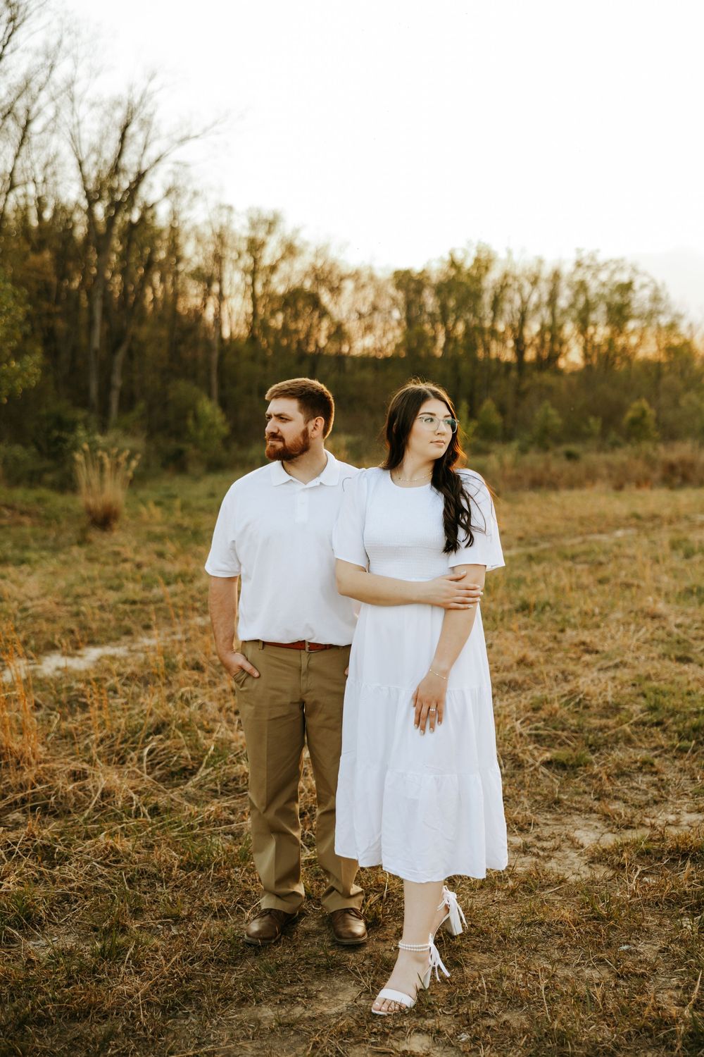 Couple stands together in a golden field at sunset wearing white outfits and embracing in warm autumn light.