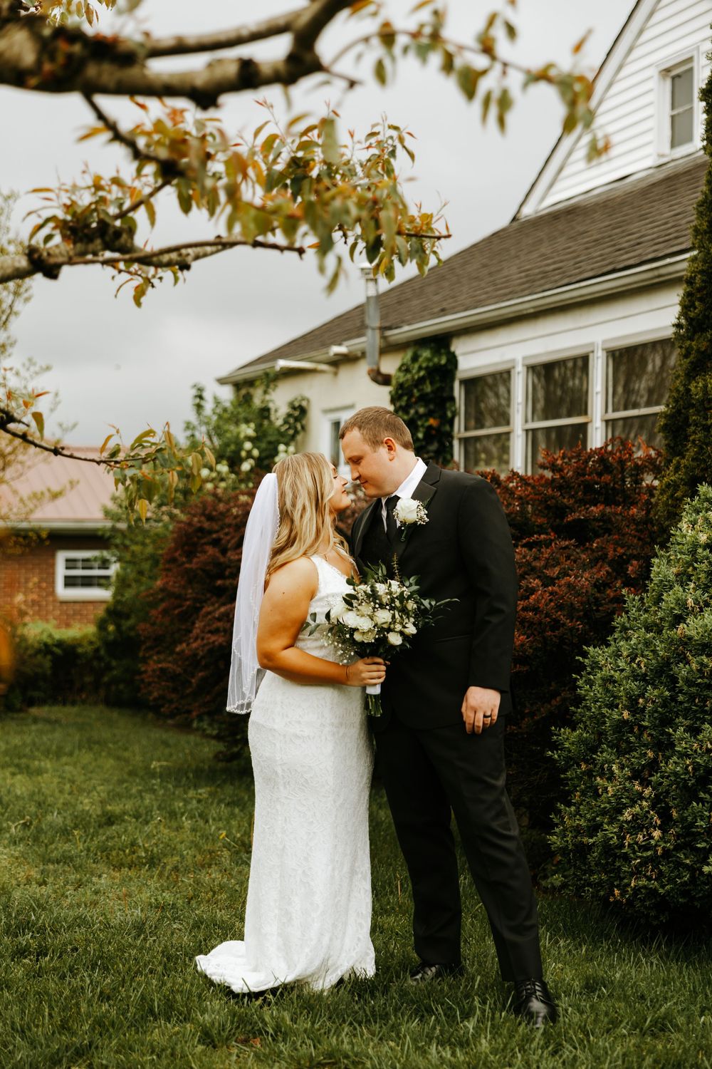 A bride and groom share a romantic moment in a garden with their wedding attire and white bouquet.