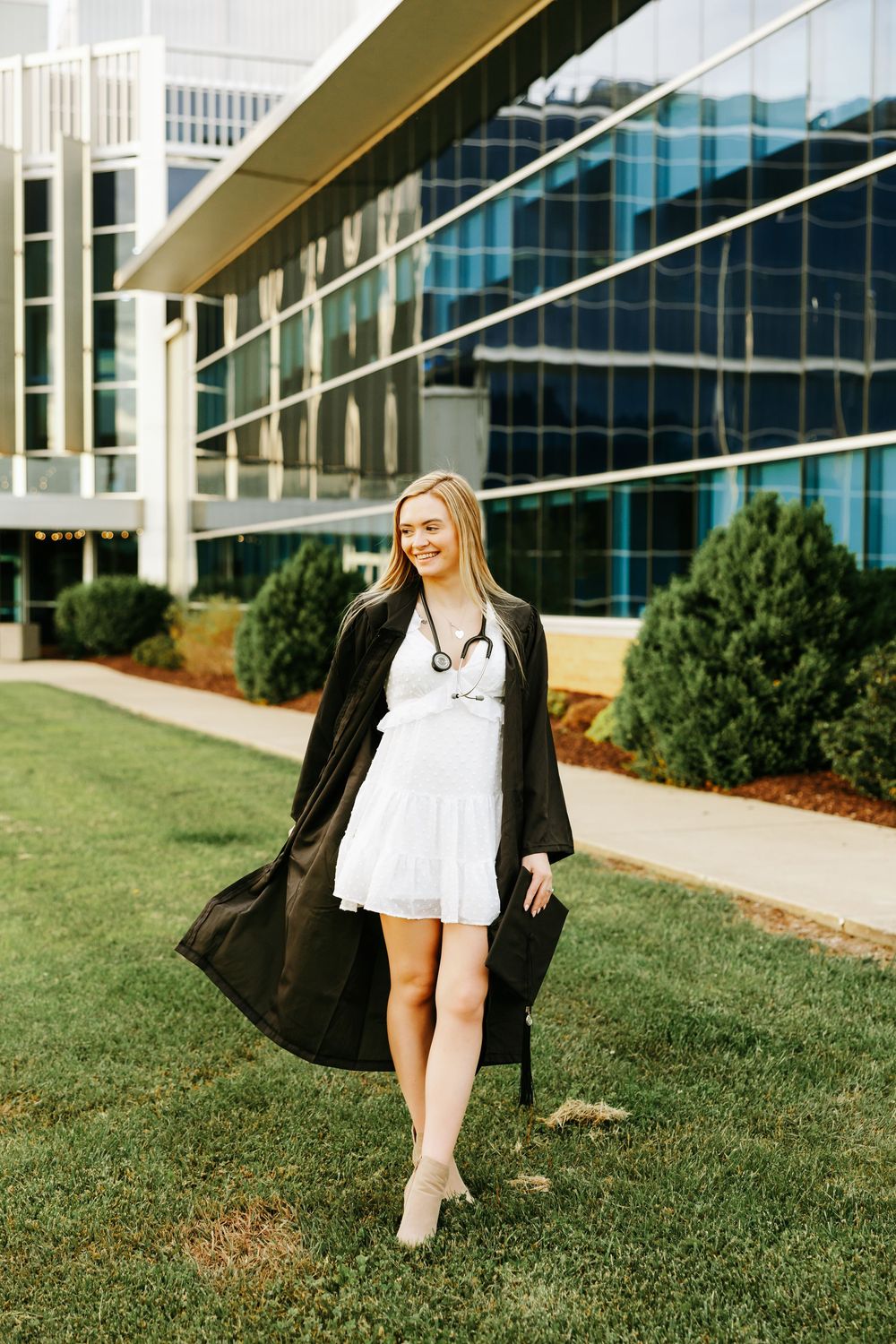Medical professional in white dress and black graduation gown stands on grass outside modern medical facility.