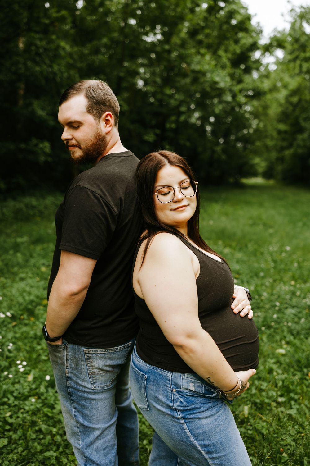A couple stands back-to-back outdoors in a grassy area surrounded by trees during a maternity photo session.