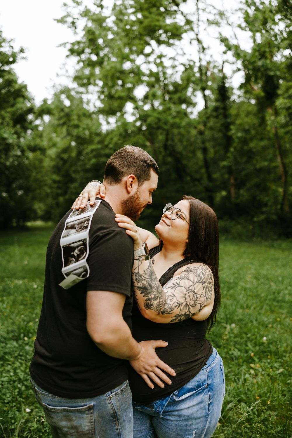 Two people share an intimate embrace while standing in a green field with trees in the background.