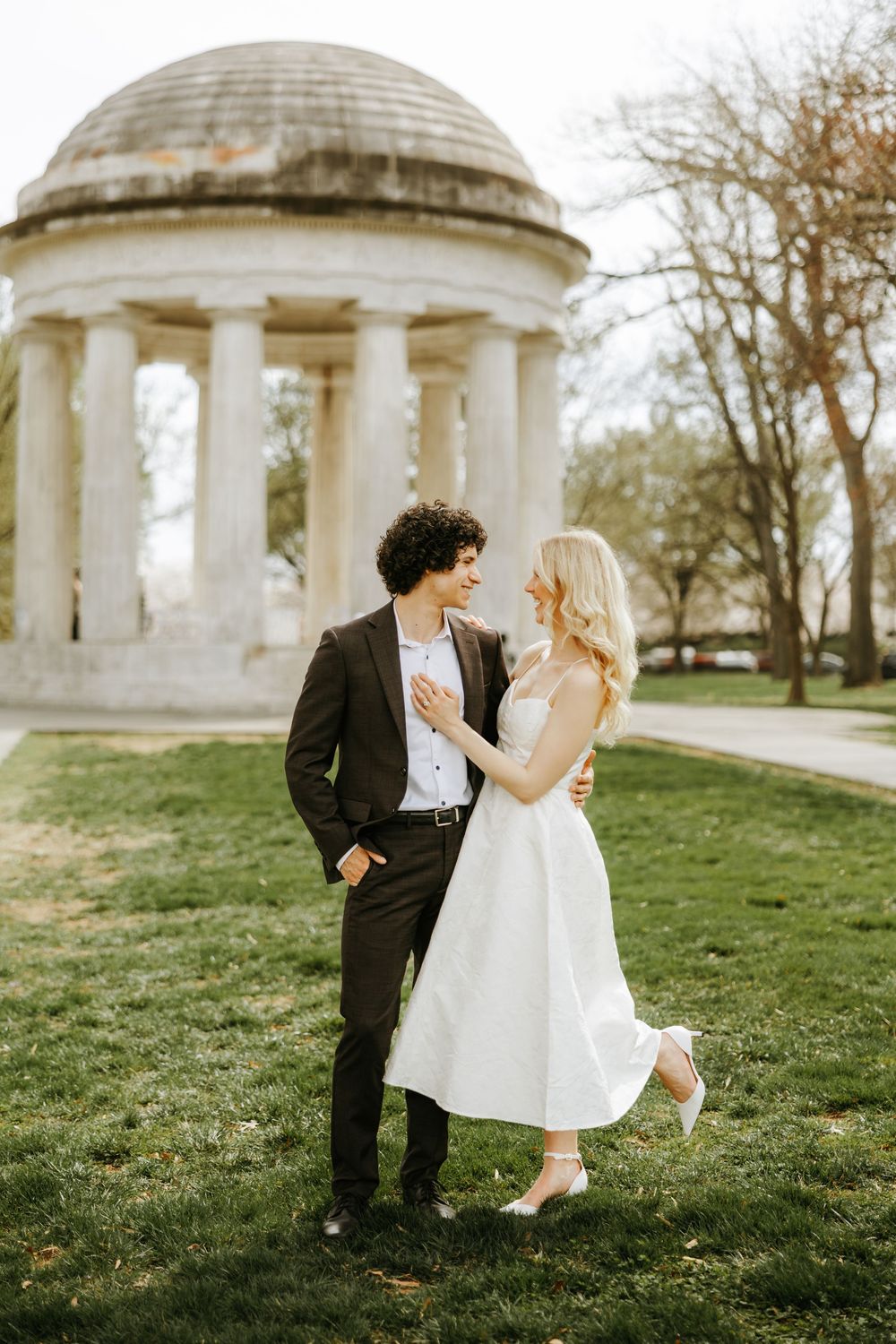 A couple in wedding attire sharing a romantic moment by a classical domed gazebo in a garden setting.