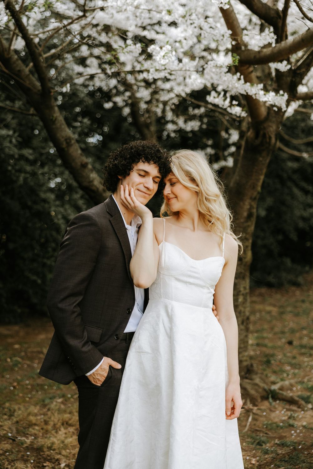 A romantic couple in formal attire shares tender moments beneath blooming cherry blossom trees in an outdoor setting.
