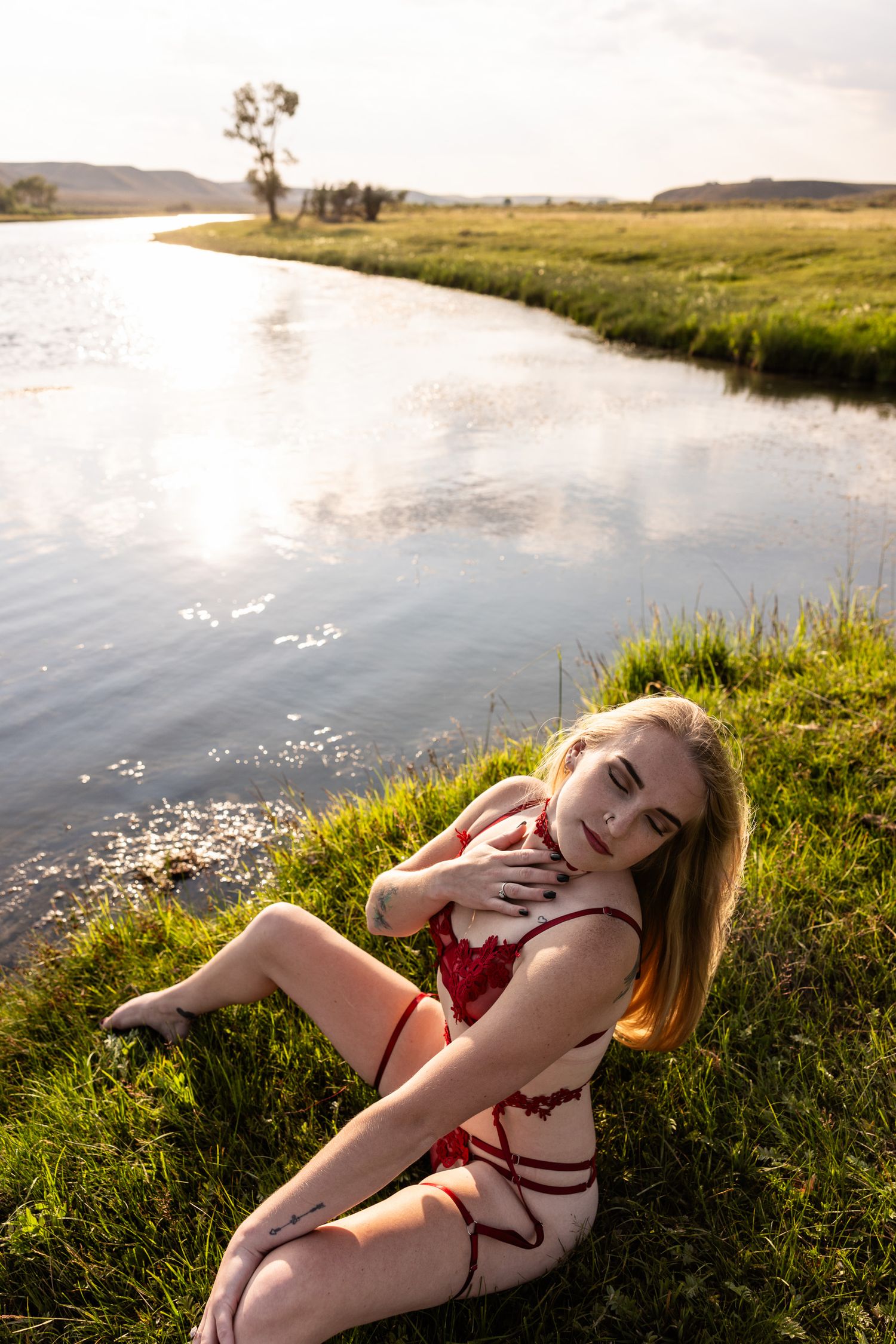 Girl in red dress poses by a serene river bank during golden hour with grassy landscape in background.