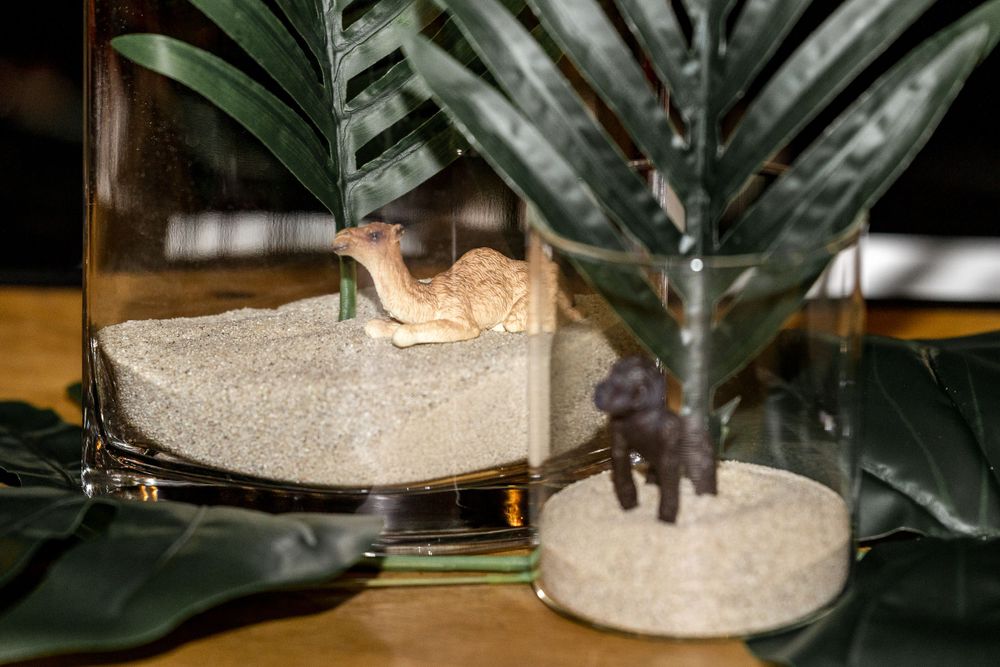 Miniature animal figurines displayed in glass terrariums with sand and tropical leaves.
