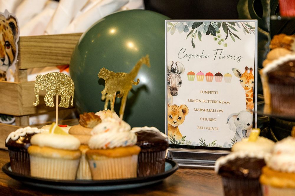 Safari-themed cupcakes with gold animal toppers next to a floral menu card and green balloon.