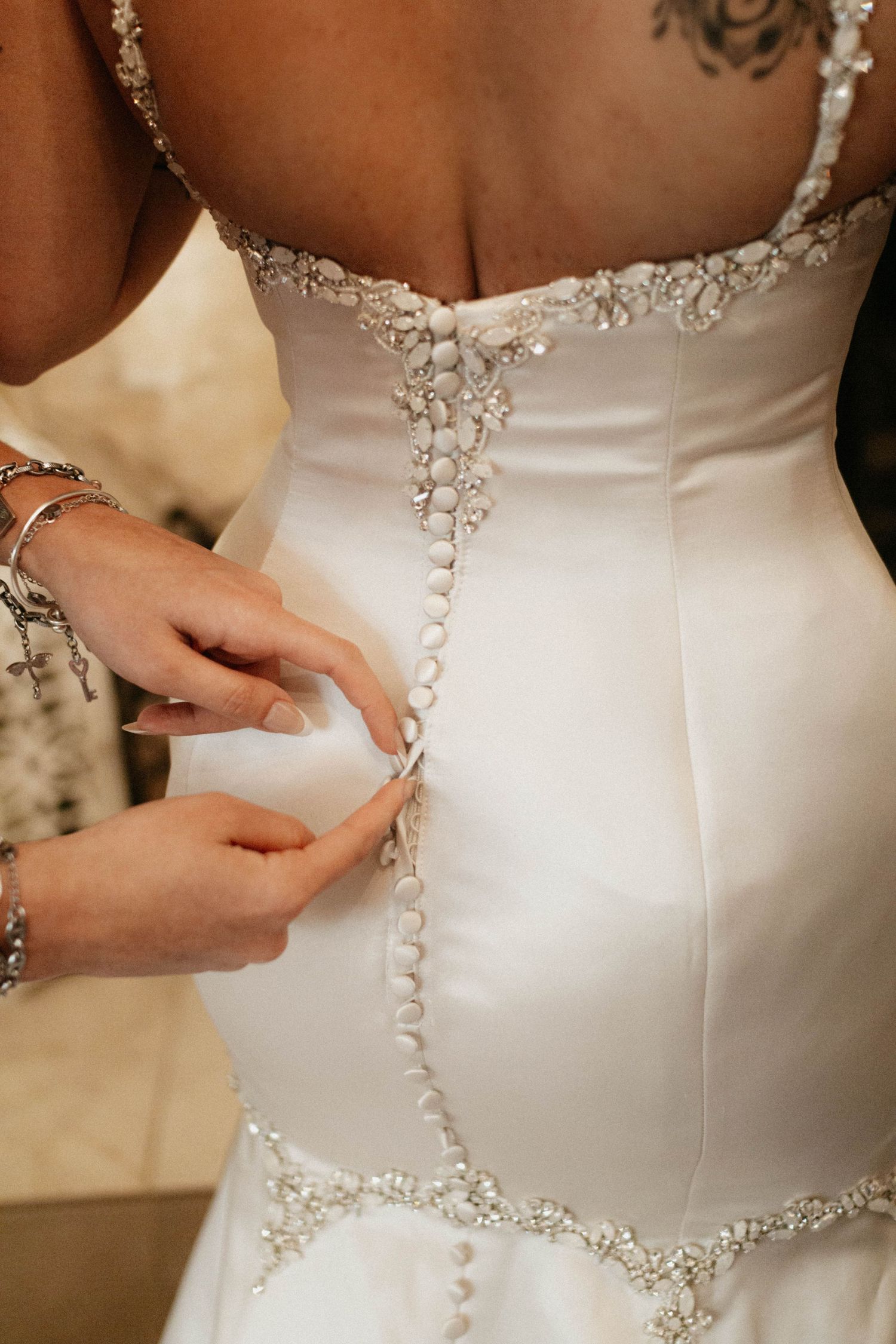 Close up of ornate buttons and beading details on the back of an elegant white satin wedding dress being fastened. Detail wedding picture
