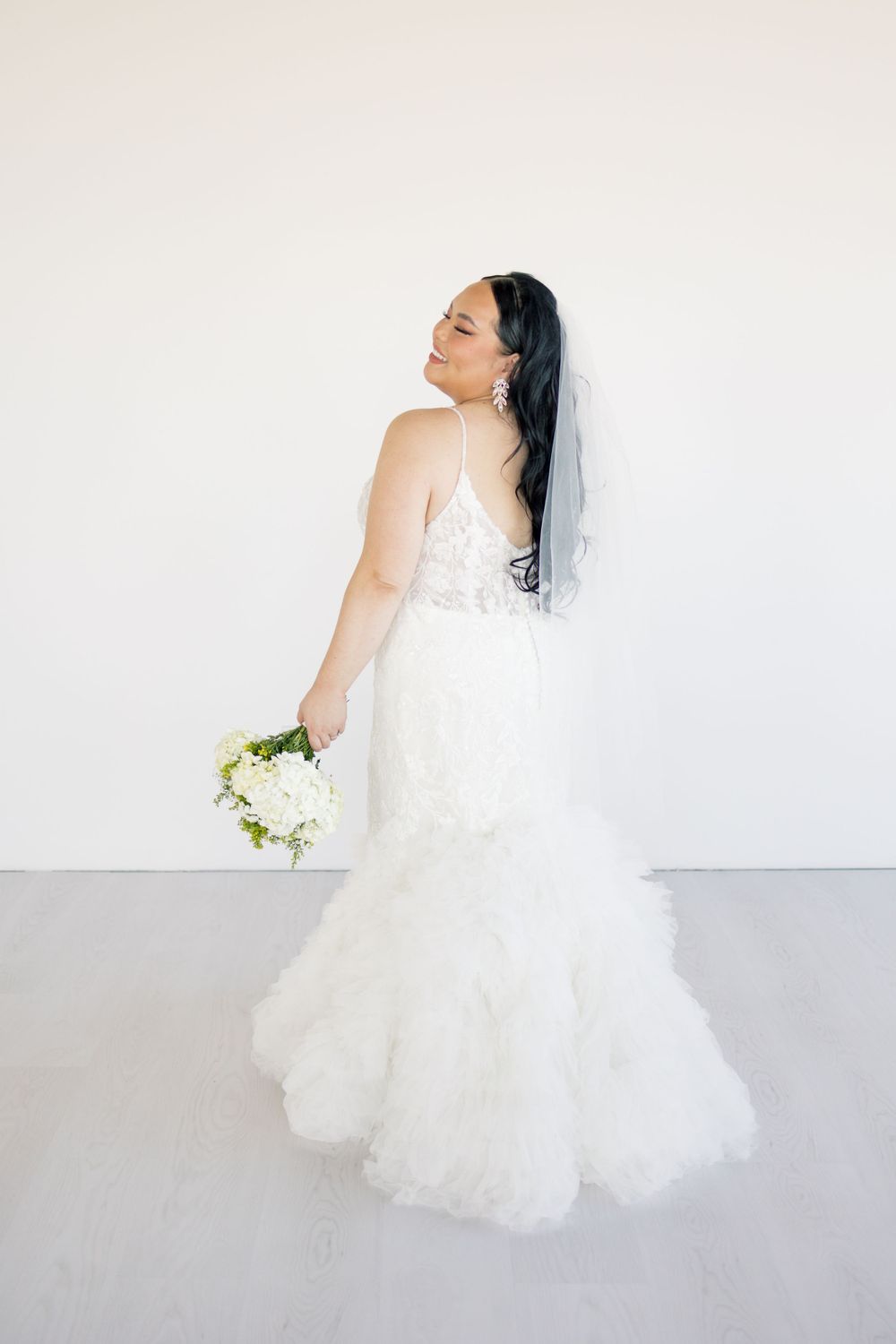 A bride in a white lace mermaid wedding gown holds a white bouquet while posing against a light backdrop.