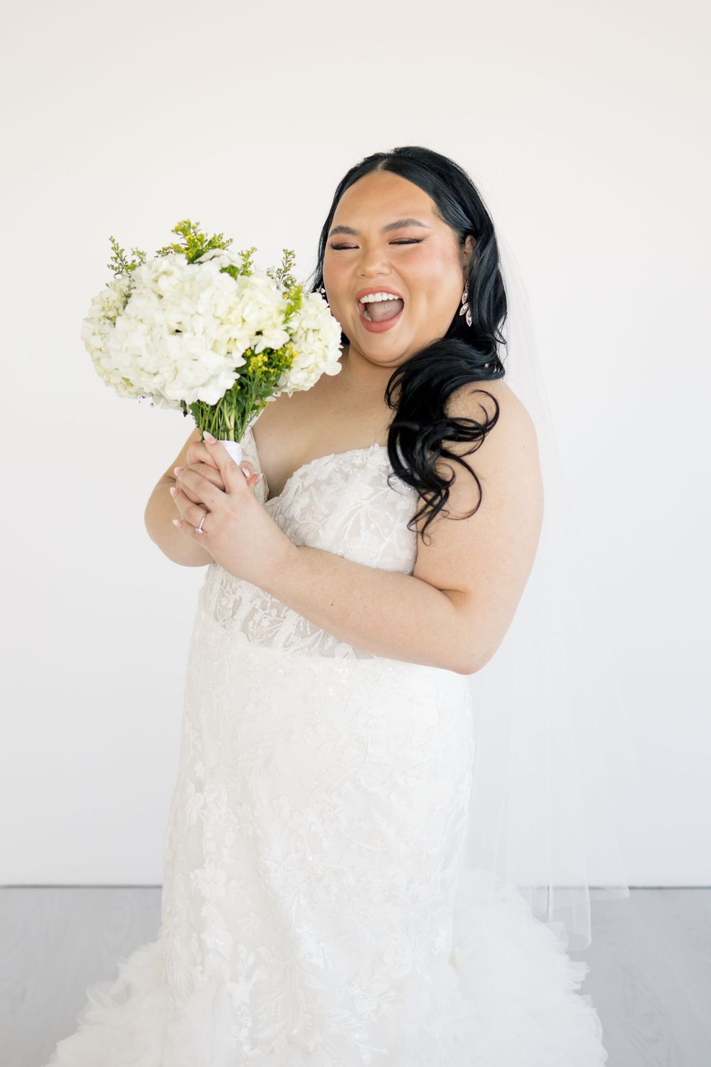 A joyful bride in a strapless wedding dress laughs while holding a white floral bouquet.