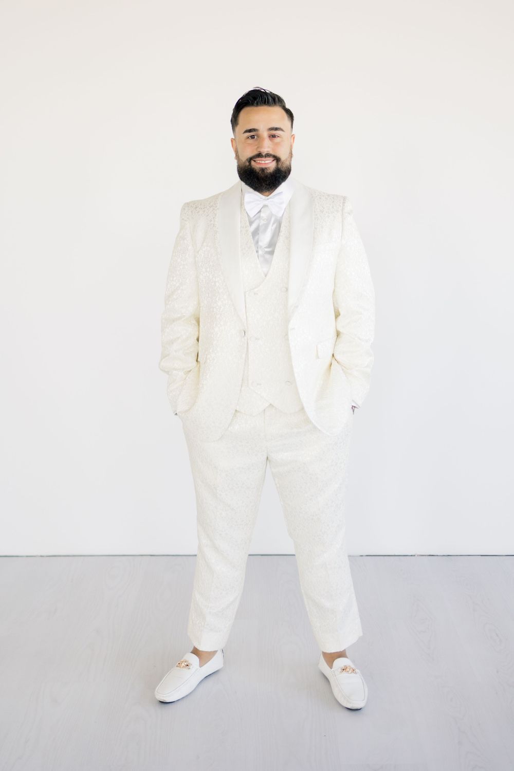 A groom in an all-white suit and white sneakers poses confidently against a light background.