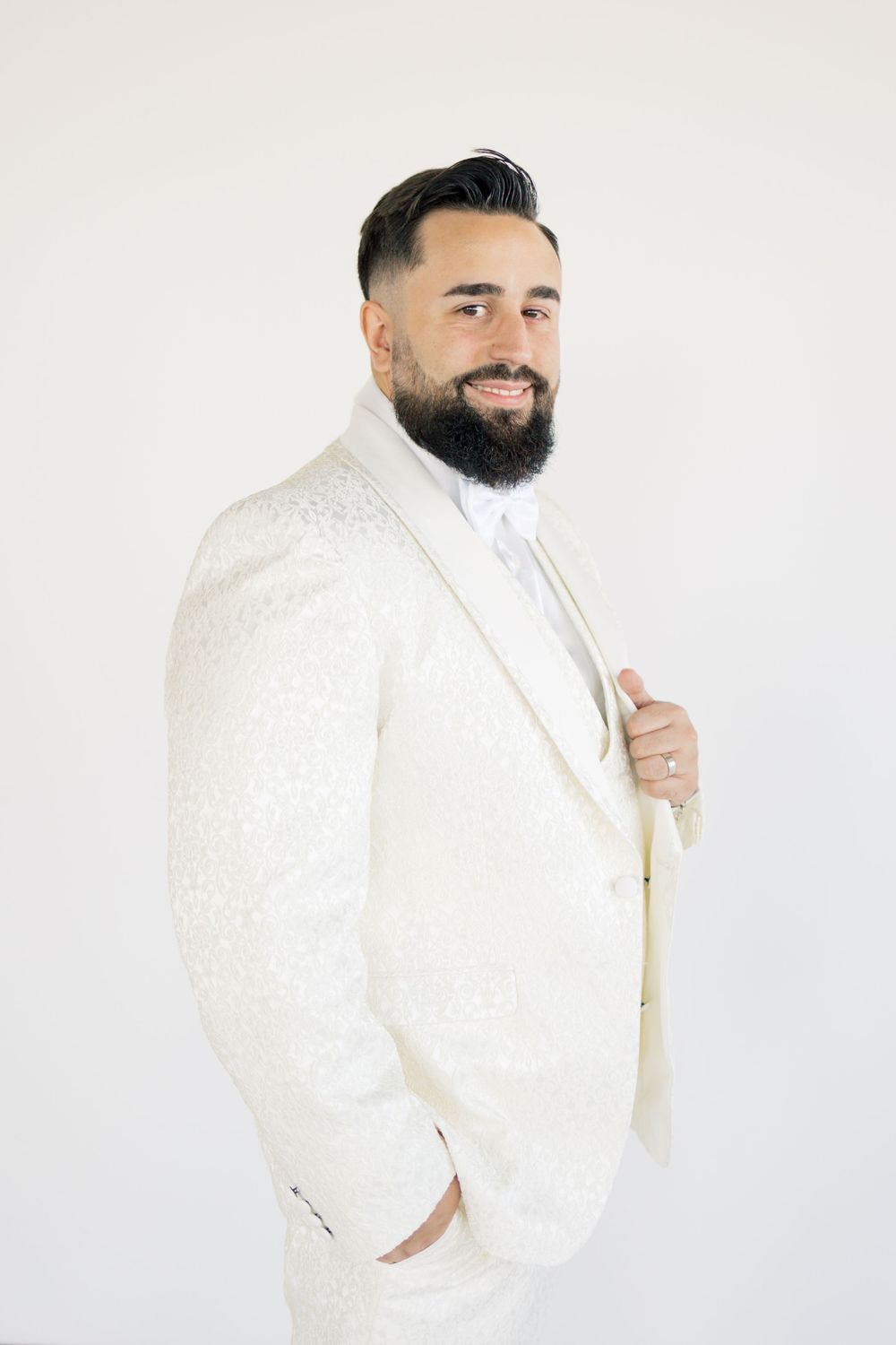 A groom in a white textured suit adjusts his lapel while smiling against a white backdrop.