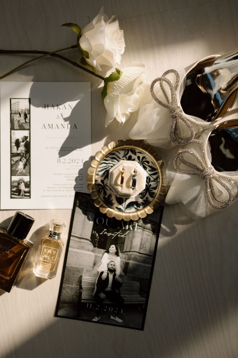Wedding details flatlay with invitation, white roses, perfume bottle and black and white photos.