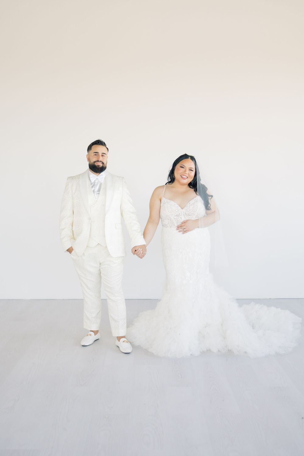 Full-length wedding portrait of couple in white against minimal background.