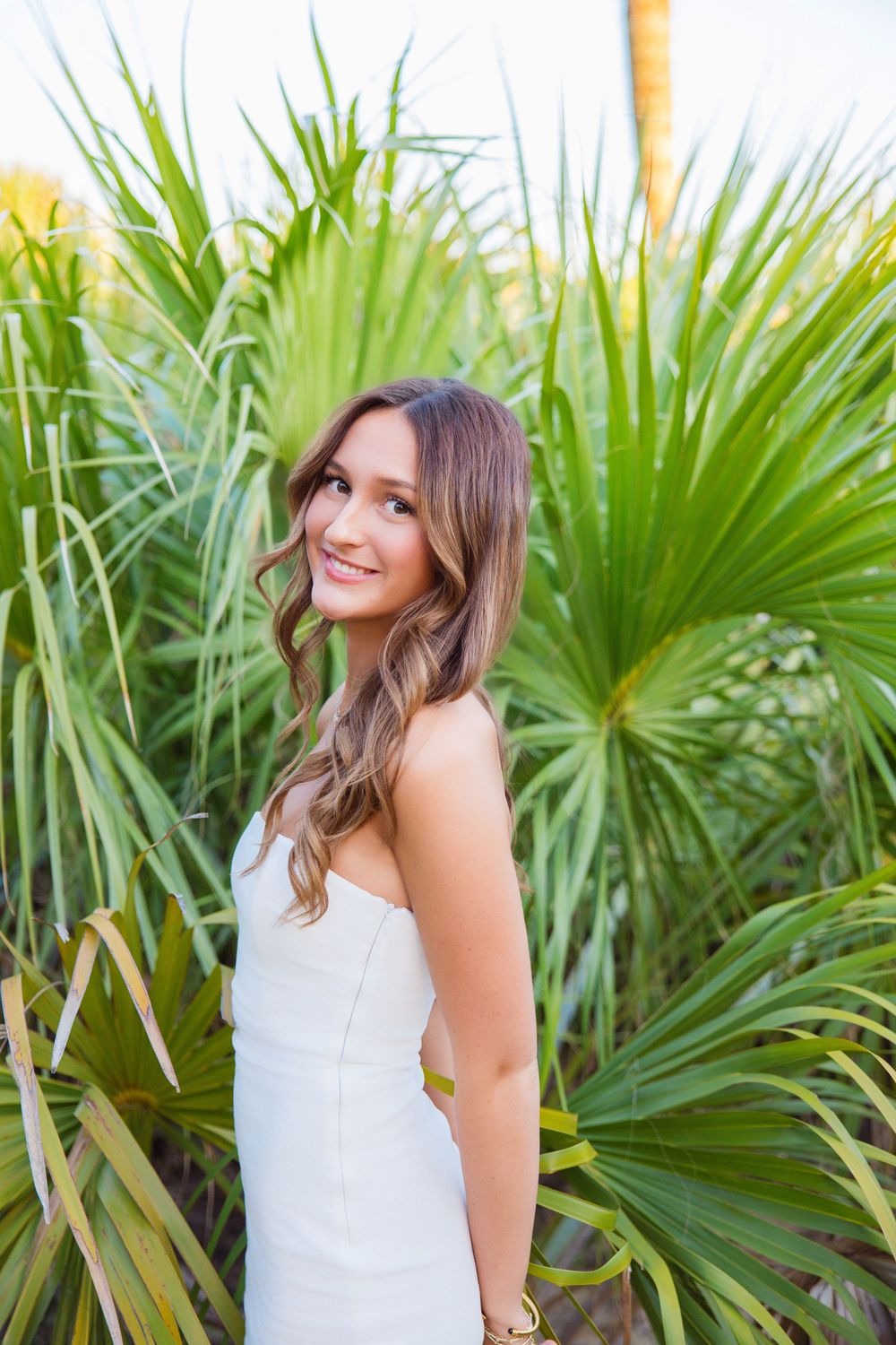 A woman in a white dress poses and smiles in front of tropical palm fronds in an outdoor setting.