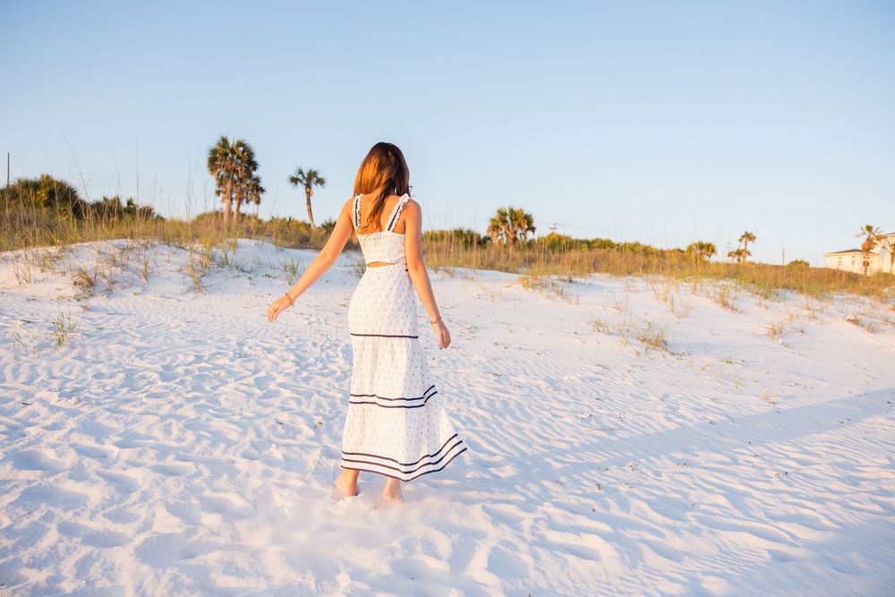 A woman in a striped white dress walks along a pristine beach during golden hour at sunset.