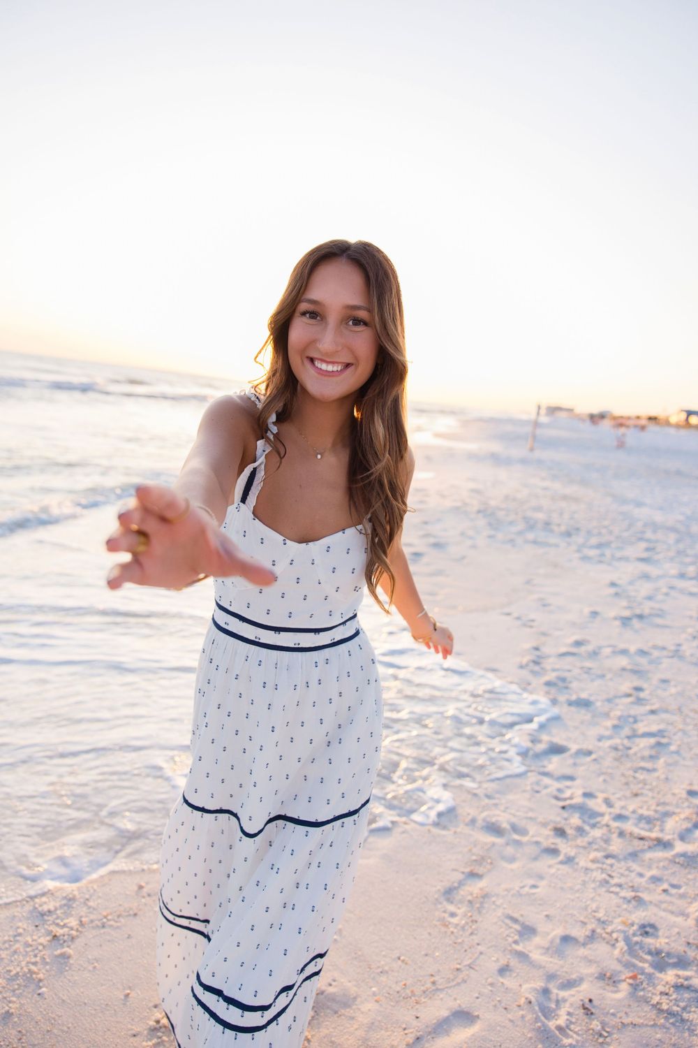 A person in a white polka dot maxi dress with navy stripes poses on a sunny beach at sunset.