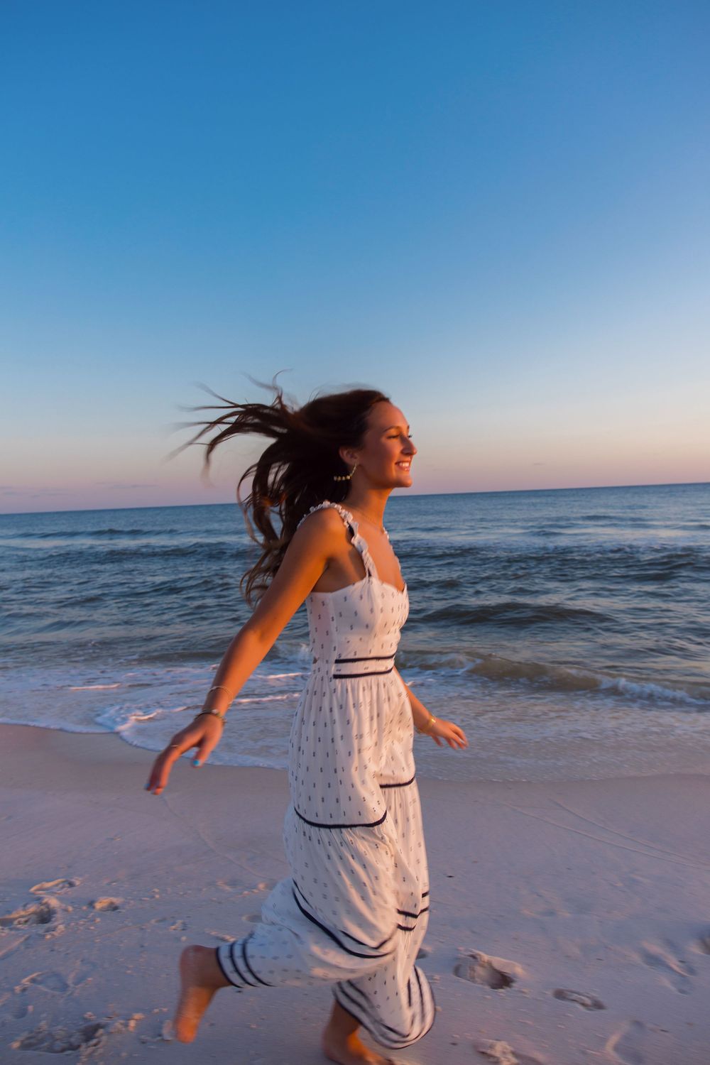A person in a white dress running joyfully along the beach at sunset with windswept hair.