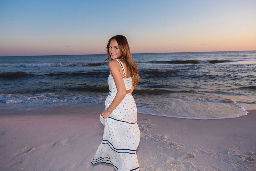A long white maxi dress flows in the ocean breeze as someone stands on a sandy beach at sunset.