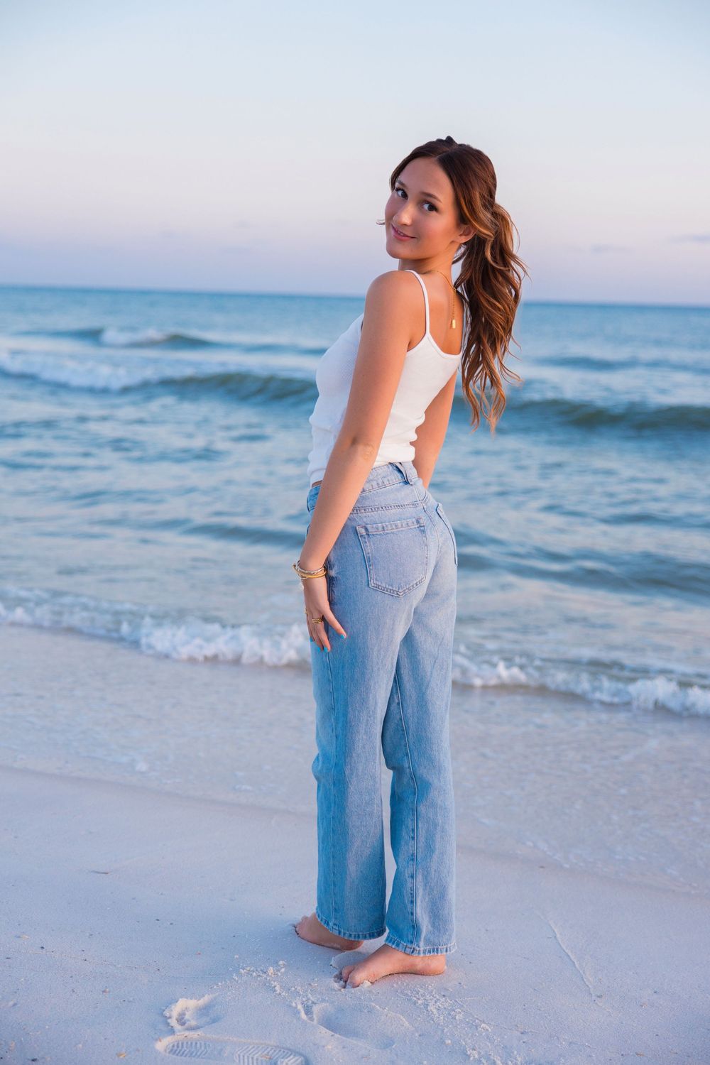 A figure poses on a sandy beach at sunset wearing a white tank top and light blue jeans beside the ocean waves.