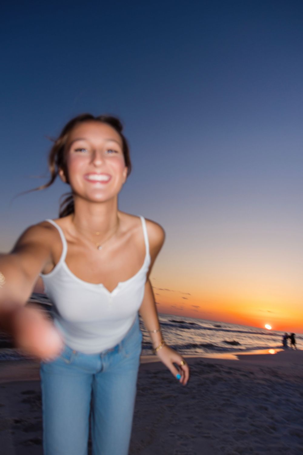 Person in white tank top and jeans enjoys a beach sunset with blurred motion effect and orange sky in background.