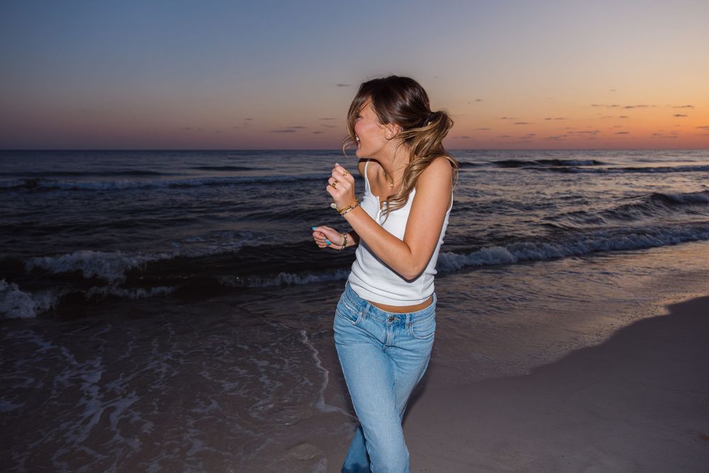 Someone enjoys a joyful moment at the beach during sunset, wearing blue jeans and a white tank top.