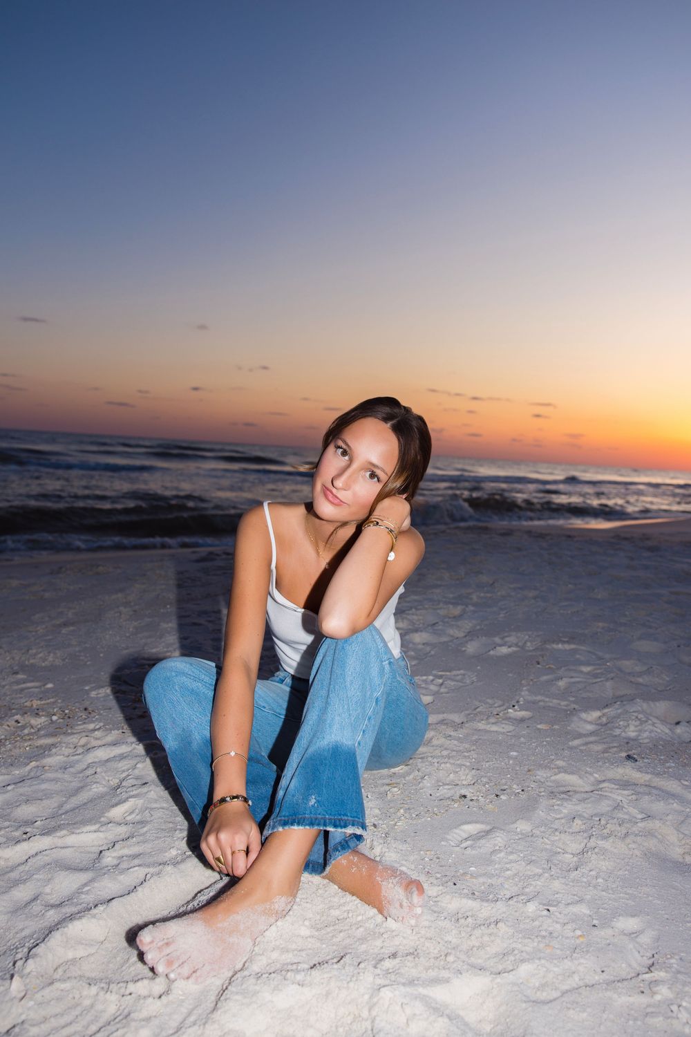A beachgoer sits in casual attire on sandy shores during a vibrant sunset over the ocean.