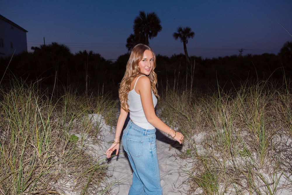 A white tank top and jeans outfit captured on a sandy beach path with palm trees at dusk.