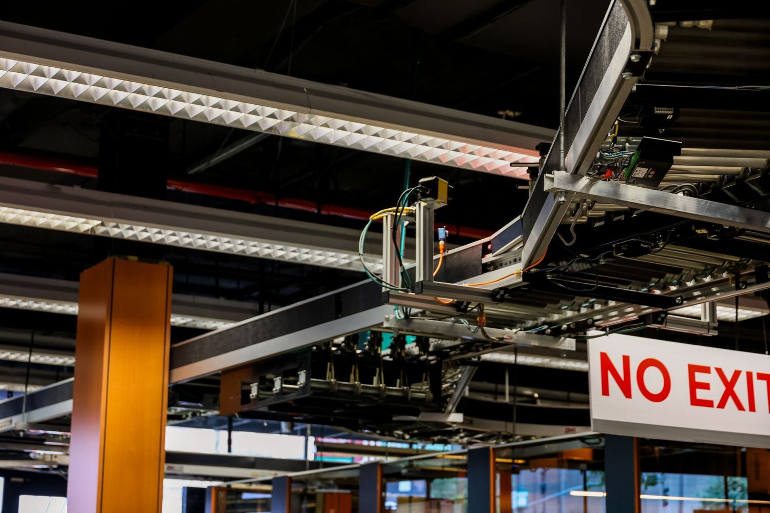 Industrial ceiling with exposed metal ducts, pipes, and a red NO EXIT sign in a commercial building interior.